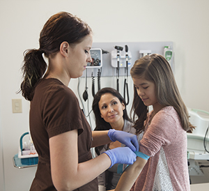Una proveedora de atención médica se prepara para tomar una muestra de sangre del brazo de una niña. La madre observa.