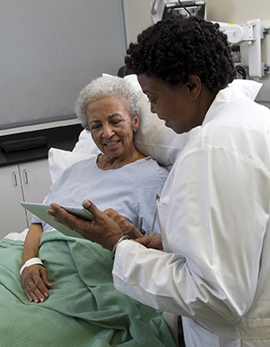 Una proveedora de atención médica sosteniendo una tableta electrónica mientras habla con una mujer en la cama de un hospital.