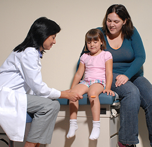 Proveedor de atención médica usando un martillo para evaluar los reflejos en la rodilla de una niña mientras una mujer los observa.